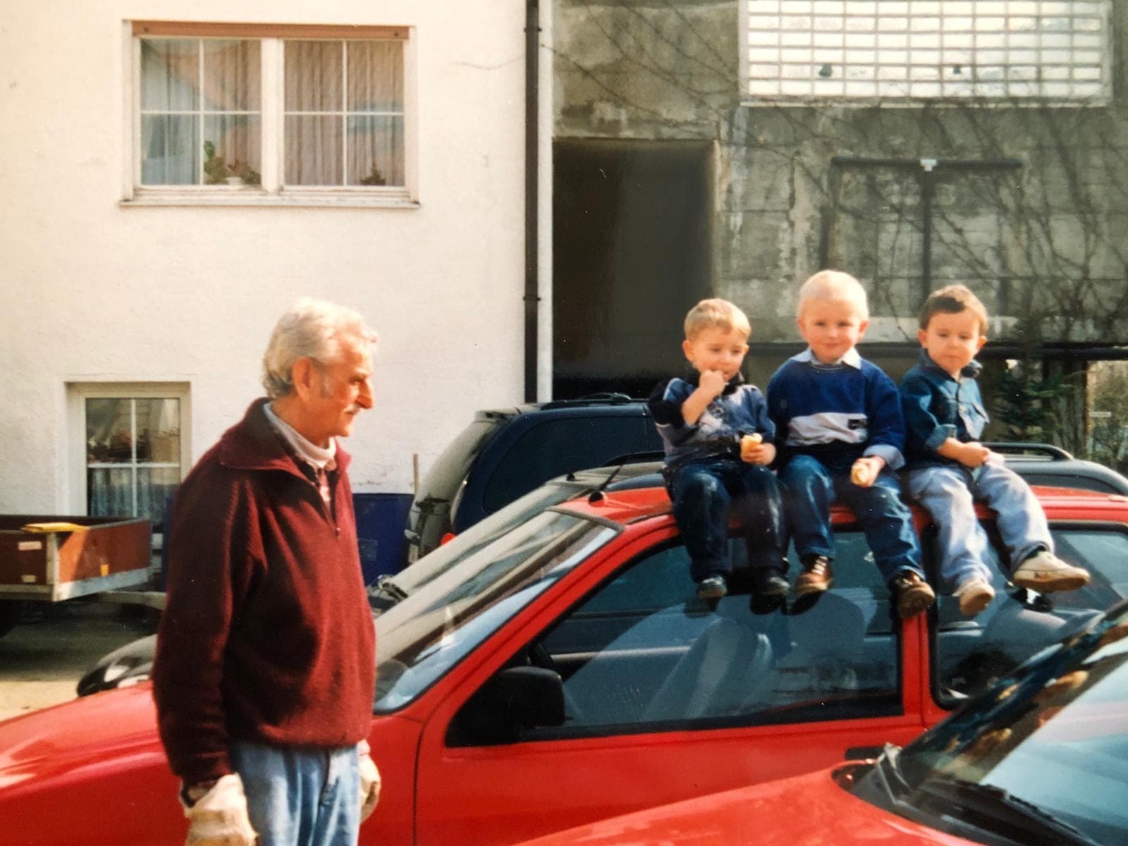 Der Großvater geht an einem roten Auto vorbei, auf dem drei Kinder sitzen